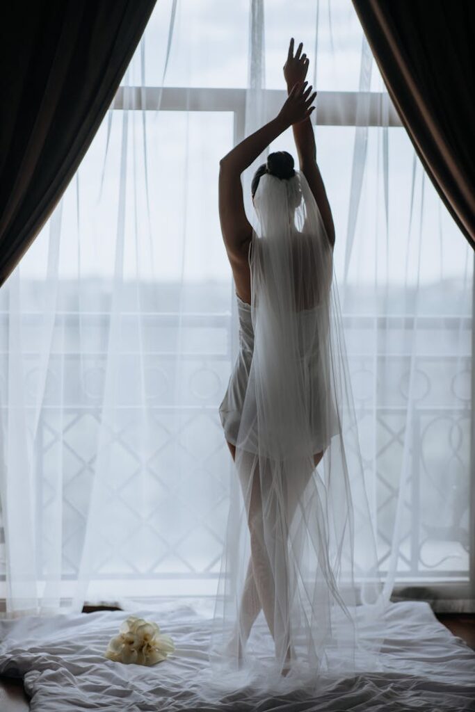 A bride poses gracefully in front of a window, draped in her wedding veil, capturing a serene morning moment.