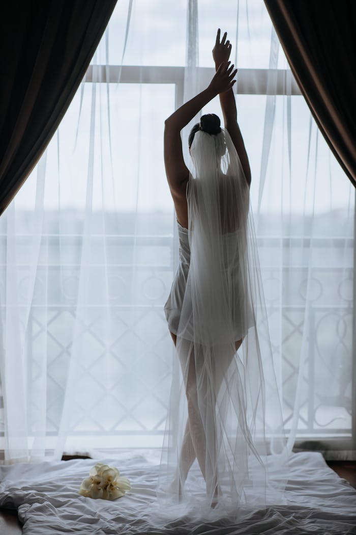 A bride poses gracefully in front of a window, draped in her wedding veil, capturing a serene morning moment.